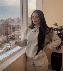a woman wearing a growth hoodie stands in front of a window holding a cup of coffee
