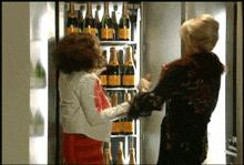 two women are standing in front of a fridge full of bottles of champagne