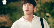 a young man wearing a white shirt with a black strap around his neck looks at the camera
