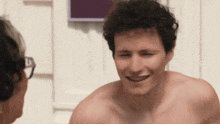 a man without a shirt is smiling at another man with glasses