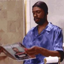 a man in a blue shirt is reading a magazine while sitting on a toilet .