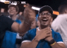 a man in a blue shirt applauds in front of a scoreboard that says goal on it