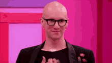 a bald man wearing glasses and earrings is standing in front of a pink wall and smiling .