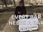 a man standing behind a sign that says " every bar needs a bar dog mind "