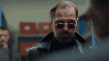 a man with a beard wearing sunglasses and a leather jacket is standing in a room .