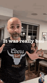 a bald man with a beard wearing a coors t-shirt is standing in a room .