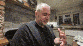 a man in a black shirt is sitting in a kitchen holding a small object in his hand .