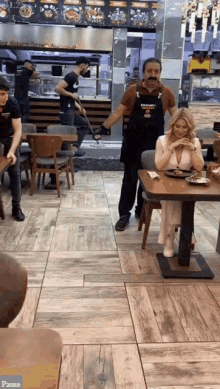a woman sits at a table in a restaurant with a man wearing an apron that says " pause "