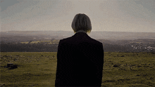 a woman in a black jacket stands on a hill overlooking a field