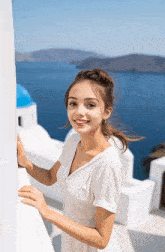 a woman in a white dress leans against a white wall and smiles