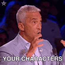 a man in a suit is speaking into a microphone and the words your characters are on the screen behind him