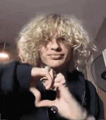 a young man with blonde curly hair is making a heart with his hands .