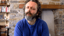 a man with a beard wearing a blue sweater is standing in front of a stone wall and a fireplace