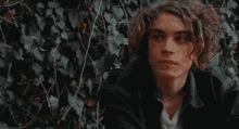a young man with curly hair is sitting in front of a leafy wall