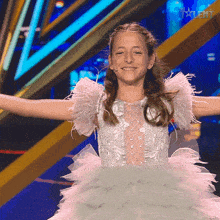 a young girl in a white dress with feathers on the sleeves is smiling on a stage