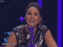 a woman in a sequined top is smiling in front of a purple microphone and has the username lialmeida05