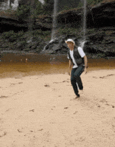 a man in a cowboy hat is squatting on a sandy beach near a waterfall