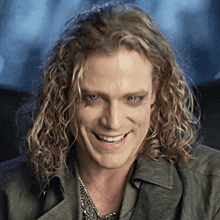 a man with long blonde curly hair and blue eyes smiles