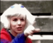 a woman wearing a white wig and a red jacket is pointing at the camera