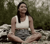 a woman in a white tank top and shorts is sitting on a rocky beach with her legs crossed .