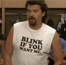 a man wearing a white tank top that says `` blink if you want me '' is standing in a locker room .