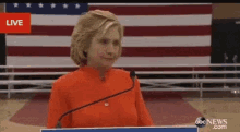 hillary clinton is giving a speech in front of an american flag while wearing an orange shirt .