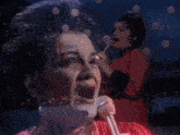 a woman in a red dress is singing into a microphone with a reflection of her face in the background