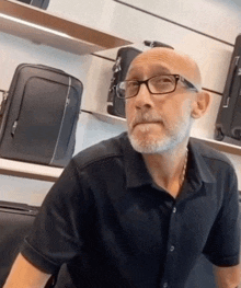 a bald man with glasses and a beard is sitting in front of a display of luggage