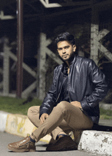 a man in a leather jacket sits on a curb with his legs crossed