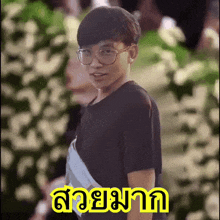 a young man wearing glasses stands in front of flowers with a yellow sticker that says ' สวย มาก '