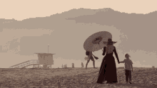 a woman in a long dress is holding an umbrella while walking on the beach with a child .