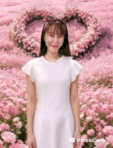 a woman in a white dress stands in front of a field of pink flowers with a heart in the background