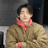 a young man wearing a jacket and a red hoodie is sitting with his arms crossed .