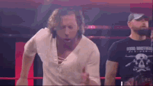 a man in a white shirt is running in a wrestling ring while another man in a black shirt stands behind him .