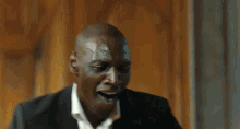 a bald man in a suit and white shirt is laughing while standing in front of a wooden wall .