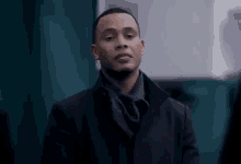a man in a black coat and scarf is standing in front of a green wall and looking at the camera .