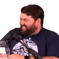 a man with a beard is laughing in front of a microphone and wearing a blue shirt with a mountain on it
