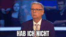 a man in a suit and tie is sitting on a stage with the words hab ich nicht written on the screen behind him .