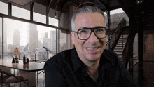 a man wearing glasses and a black shirt smiles in front of a large window