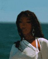 a woman wearing earrings and a white shirt stands in front of the ocean