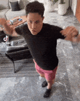 a man in a black shirt and pink shorts is dancing in a living room with a tik tok sticker on the corner