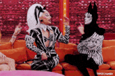 two drag queens are sitting on a couch and one of them is wearing a black and white outfit