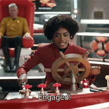 a woman in a red jacket is holding a steering wheel and the word engaged is on the screen