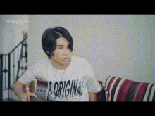 a young man is sitting on a couch holding a guitar and wearing a t-shirt that says originals .