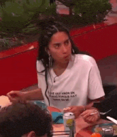 a woman in a white t-shirt is sitting at a table with a can of beer .