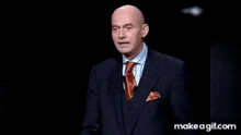 a bald man in a suit and tie is saluting while standing on a stage .
