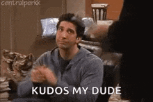 a man is sitting at a table with his hands folded in front of him and a sign that says kudos my dude .
