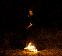 a woman in a black dress stands in front of a fire in the dark