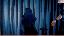 a man stands in front of a blue curtain behind a gaming chair that says nzxt on the back