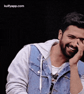 a man with a beard and a denim jacket is laughing while covering his face with his hand .